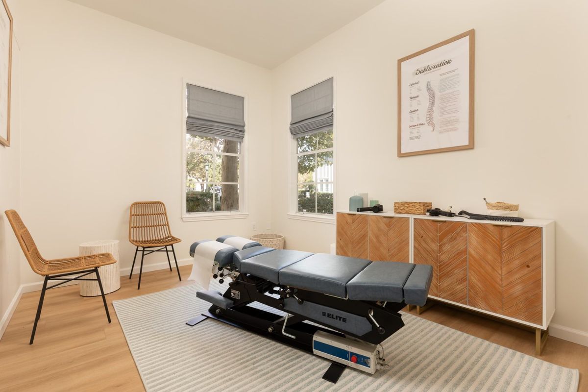 A bright Vitality treatment room with blush armchairs, a patterned rug and an adjusting table