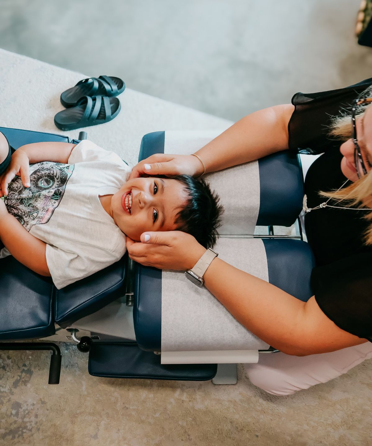 A laughing boy lies on an adjusting table while a chiropractor gently holds his head