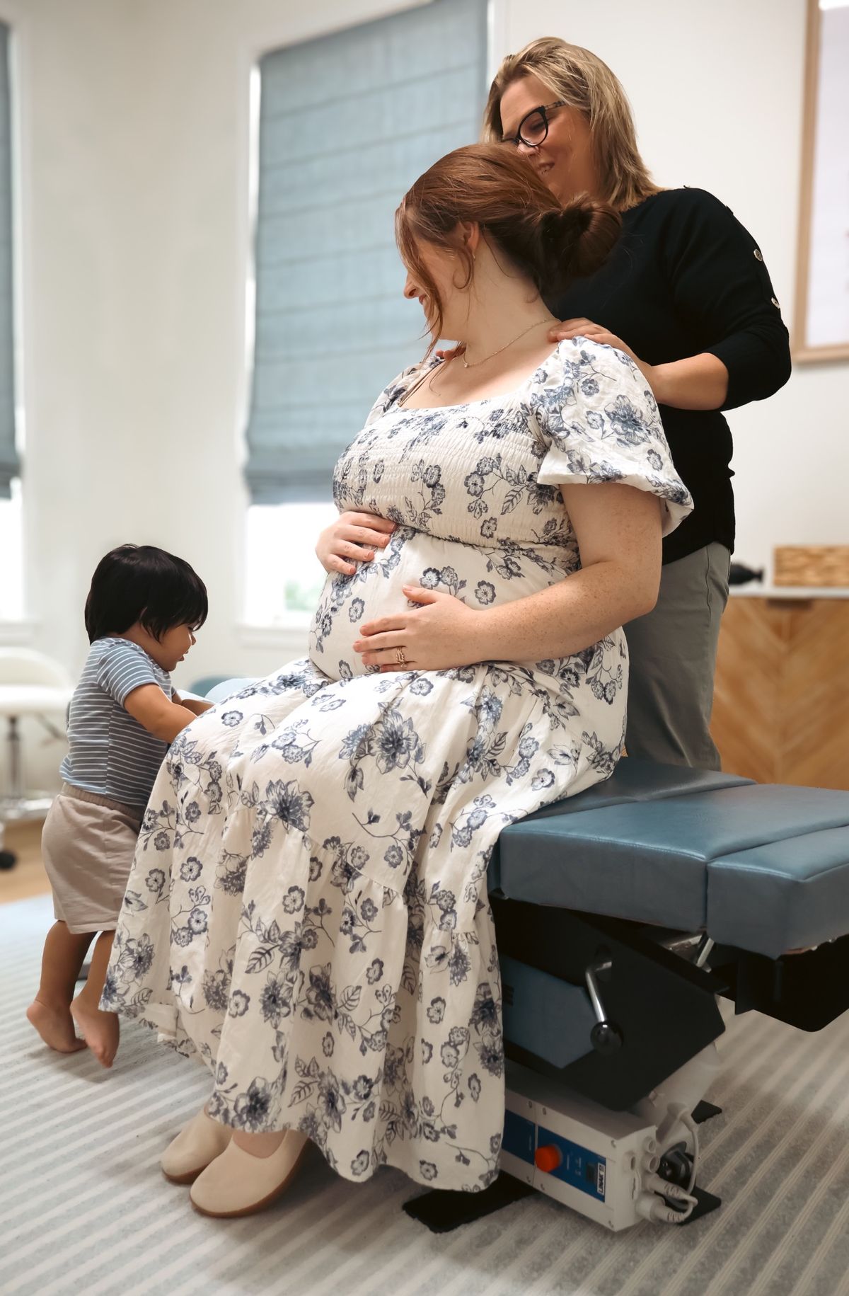 A smiling pregnant mom sits on an adjusting table with her hands on her belly while a chiropractor stands behind her and her toddler plays nearby