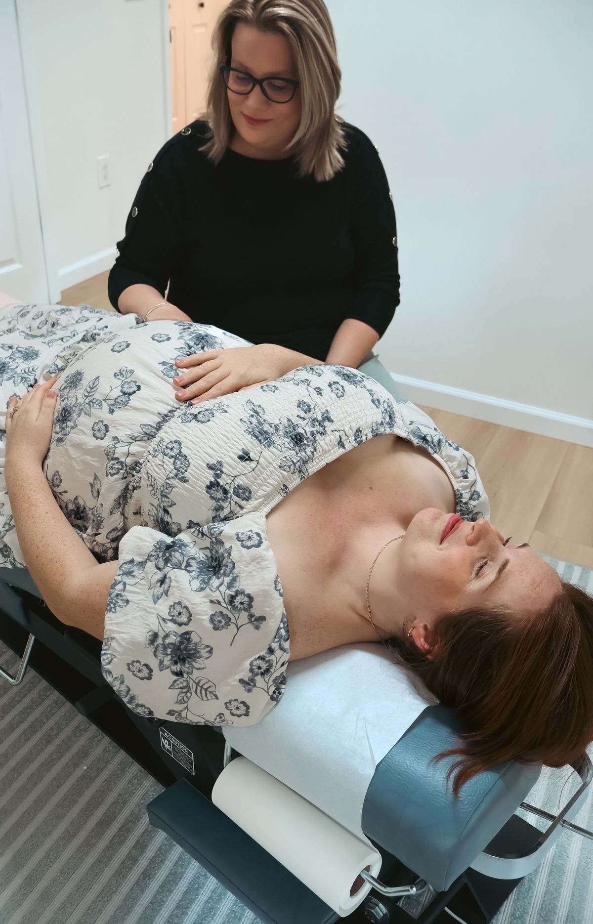 A pregnant mom rests face up on a chiropractic table, eyes closed, while a chiropractor rests a hand on her belly